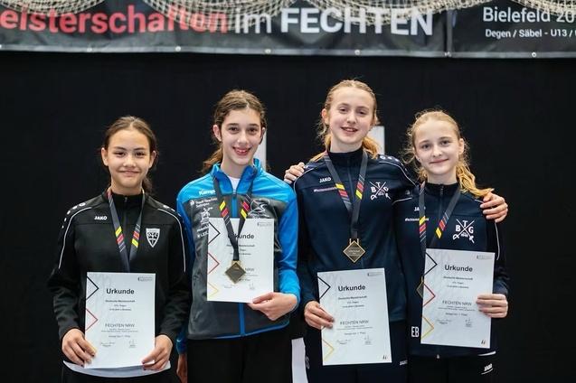Vier fechtende M&auml;dchen halten ihre Urkunden in der Hand, l&auml;chelnd, mit Medaillen um den Hals, im Hintergrund ein Banner.
