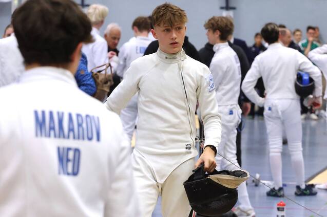 Junger Fechter in wei&szlig;er Ausr&uuml;stung h&auml;lt seine Maske in einer Turnhalle mit anderen Sportlern im Hintergrund.