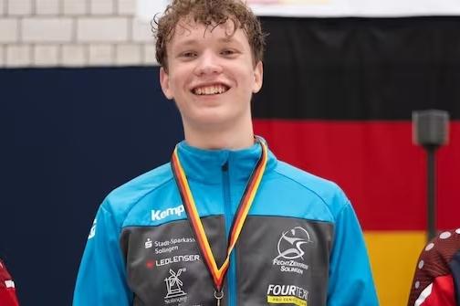 Junger Sportler mit Medaille, l&auml;chelt vor einer deutschen Flagge in einem Wettkampfumfeld.
