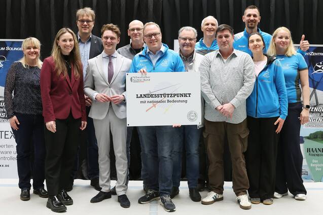 Gruppenfoto von zehn Personen, die ein Schild mit der Aufschrift &bdquo;LANDESST&Uuml;TZPUNKT mit sportlicher Bedeutung&ldquo; halten.