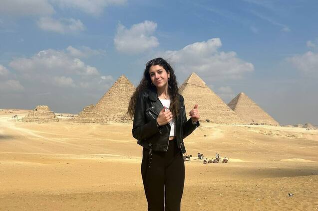 Junge Frau in Lederjacke steht im Vordergrund vor den Pyramiden von Gizeh bei klarem Himmel und Sandboden.