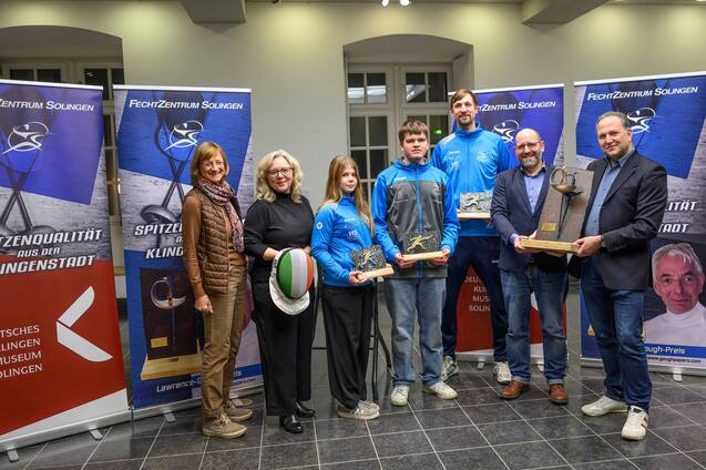 Gruppenszene mit sechs Personen vor Banner, die Pokale und Auszeichnungen beim Fechtsport zeigen.