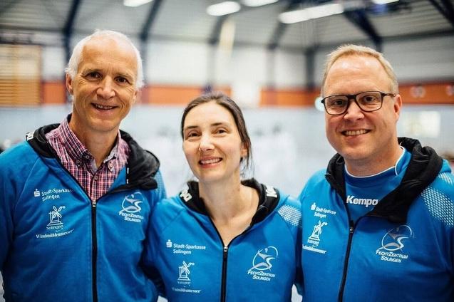 Drei Personen in blauen Sportjacken posieren l&auml;chelnd in einer Sporthalle. Hintergrund zeigt Trainingsger&auml;te.
