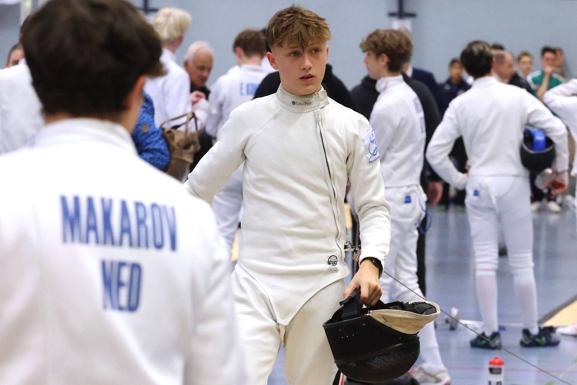 Junger Fechter in weißer Ausrüstung hält seine Maske in einer Turnhalle mit anderen Sportlern im Hintergrund.