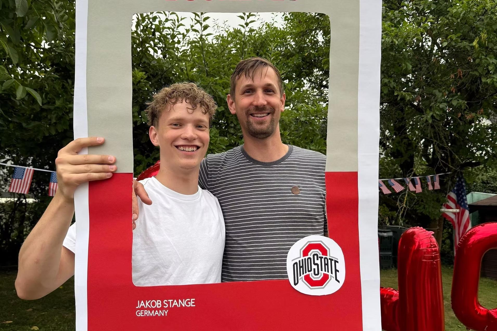 Zwei M&auml;nner posieren l&auml;chelnd vor einem gro&szlig;en, rahmenartigen Schild mit der Aufschrift &bdquo;BUCKEYES&ldquo;.