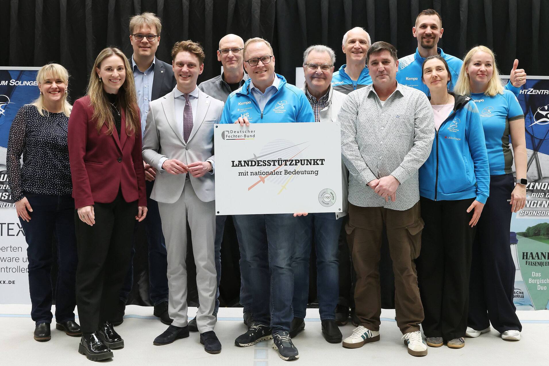 Gruppenfoto von zehn Personen, die ein Schild mit der Aufschrift &bdquo;LANDESST&Uuml;TZPUNKT mit sportlicher Bedeutung&ldquo; halten.