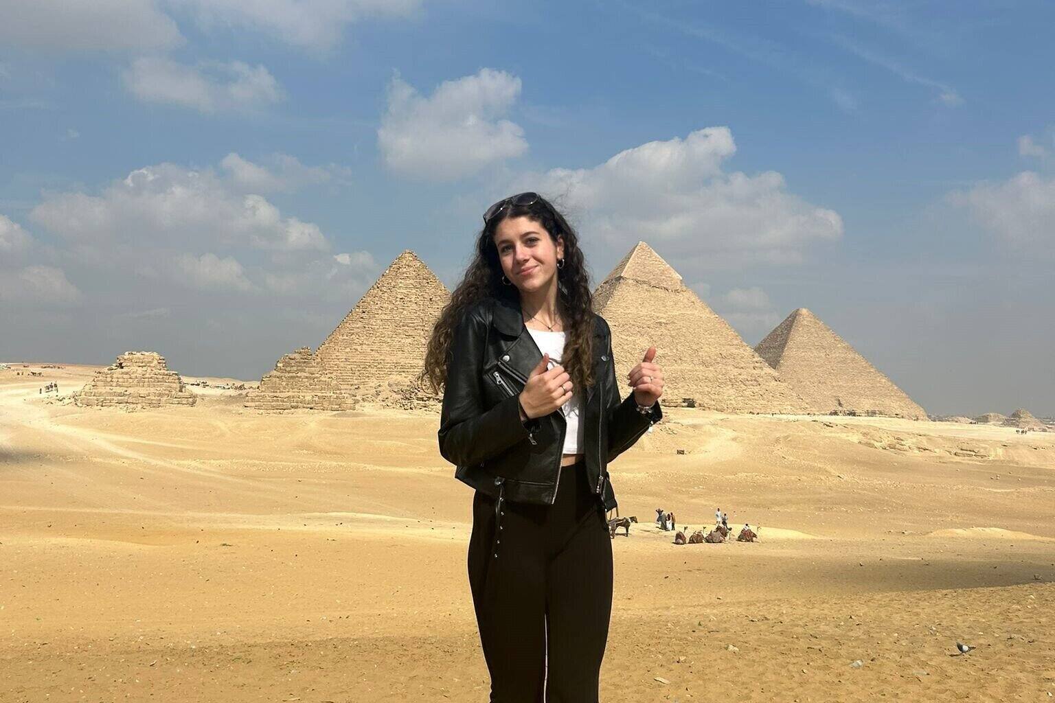 Junge Frau in Lederjacke steht im Vordergrund vor den Pyramiden von Gizeh bei klarem Himmel und Sandboden.