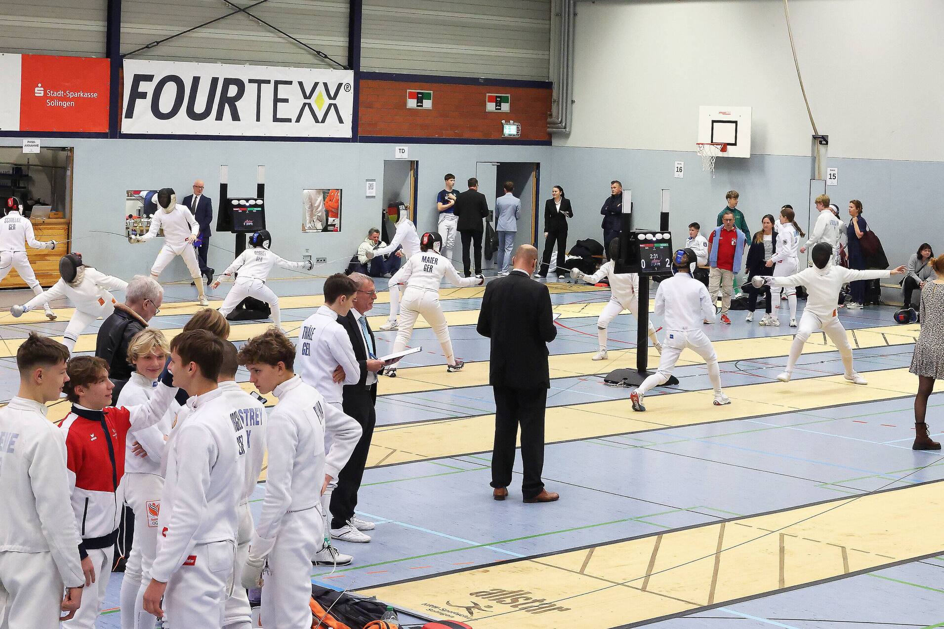 Fencers in white uniforms train auf mehreren Matten in einer Sporthalle, Zuschauer beobachten die Aktivit&auml;ten im Hintergrund.