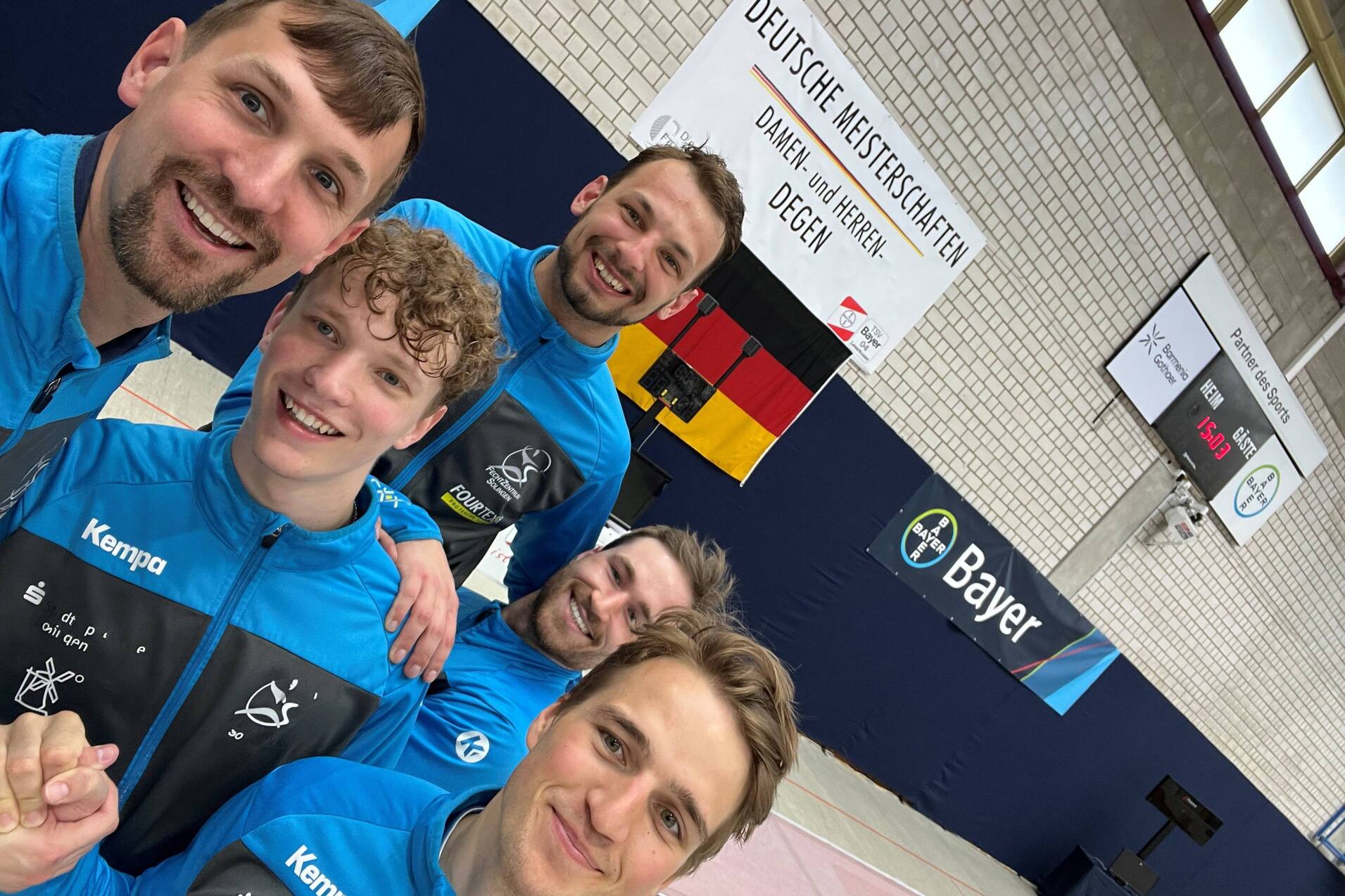 Fünf Männer in blauen Trainingsjacken lächeln in die Kamera vor einer Wettkampfstätte mit Banner und Flaggen.