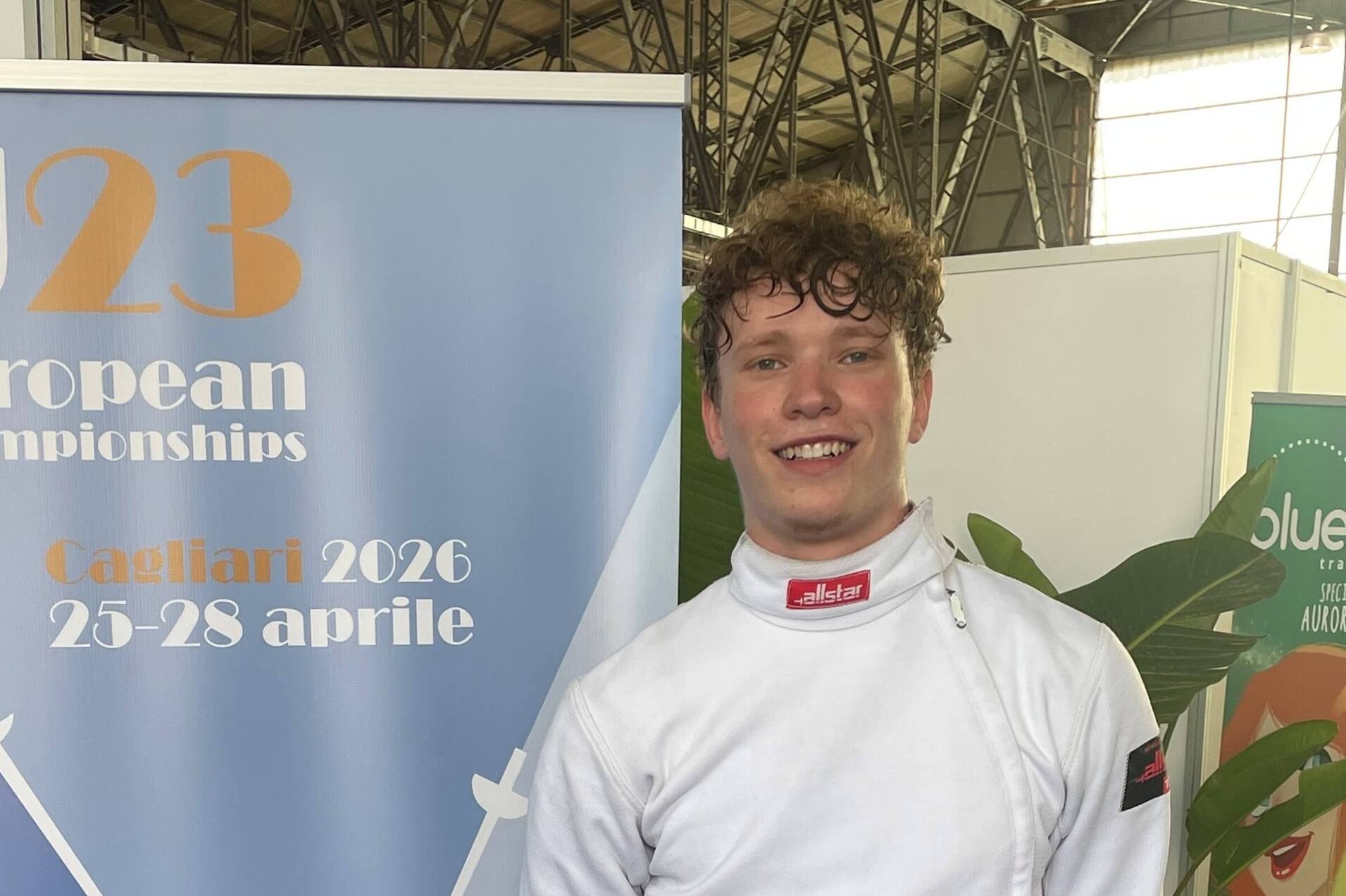 Junger Fechter in Wettkampfuniform steht neben einem Banner der U23 Europameisterschaften 2026 in Cagliari.