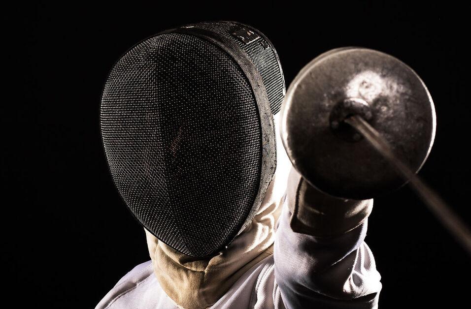 Fencer in protective gear, holding a foil with an outstretched arm against a dark background.