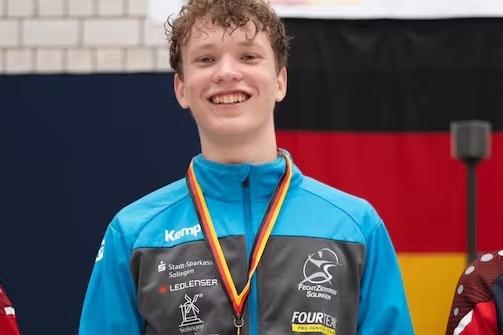 Junger Sportler mit Medaille, lächelt vor einer deutschen Flagge in einem Wettkampfumfeld.