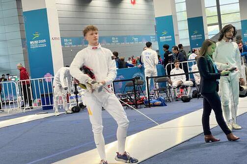 Fencer in Wettkampfausrüstung mit Maske und Degen auf einer blauem Fläche, umgeben von anderen Teilnehmern und Zuschauern.