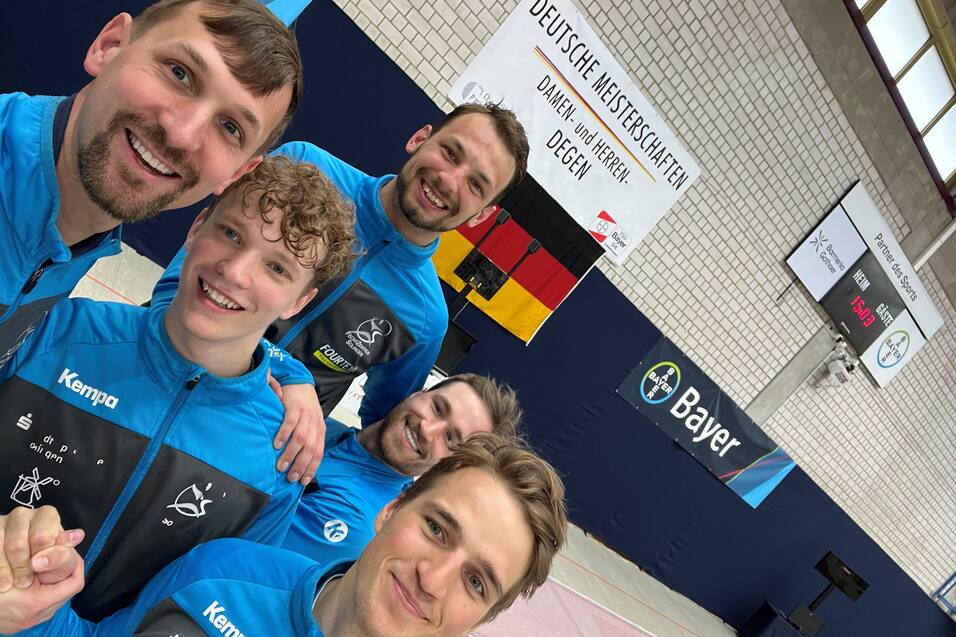 Fünf Männer in blauen Trainingsjacken lächeln in die Kamera vor einer Wettkampfstätte mit Banner und Flaggen.