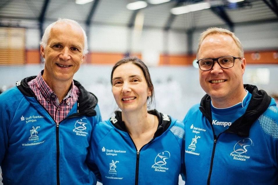 Drei Personen in blauen Sportjacken posieren lächelnd in einer Sporthalle. Hintergrund zeigt Trainingsgeräte.