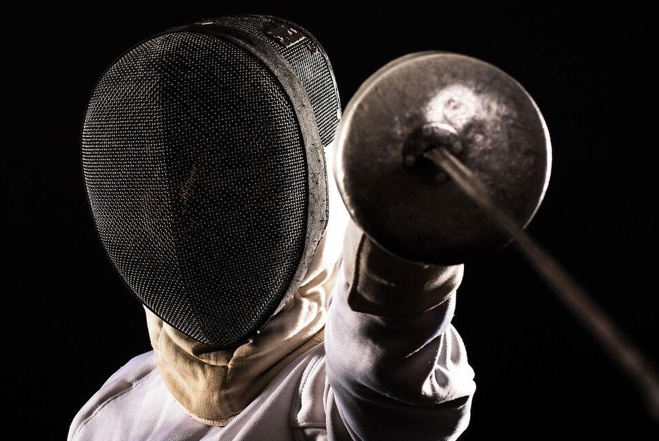Fencer in protective gear, holding a foil with an outstretched arm against a dark background.