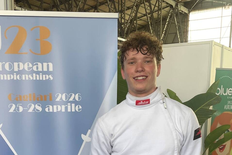 Junger Fechter in Wettkampfuniform steht neben einem Banner der U23 Europameisterschaften 2026 in Cagliari.