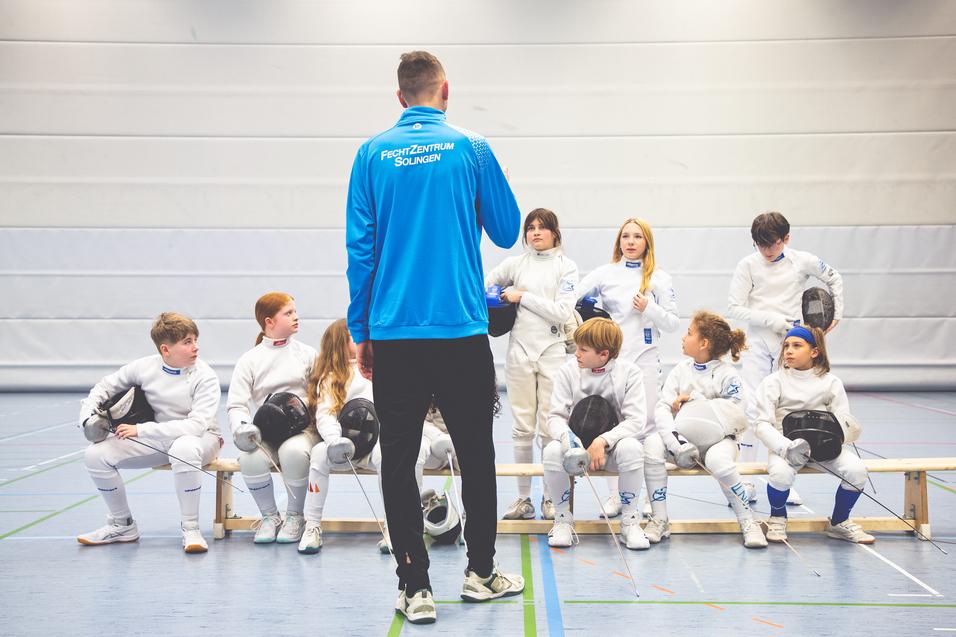 Trainer spricht mit einer Gruppe junger Fechter in Wettkampfanz&uuml;gen in einer Sporthalle.