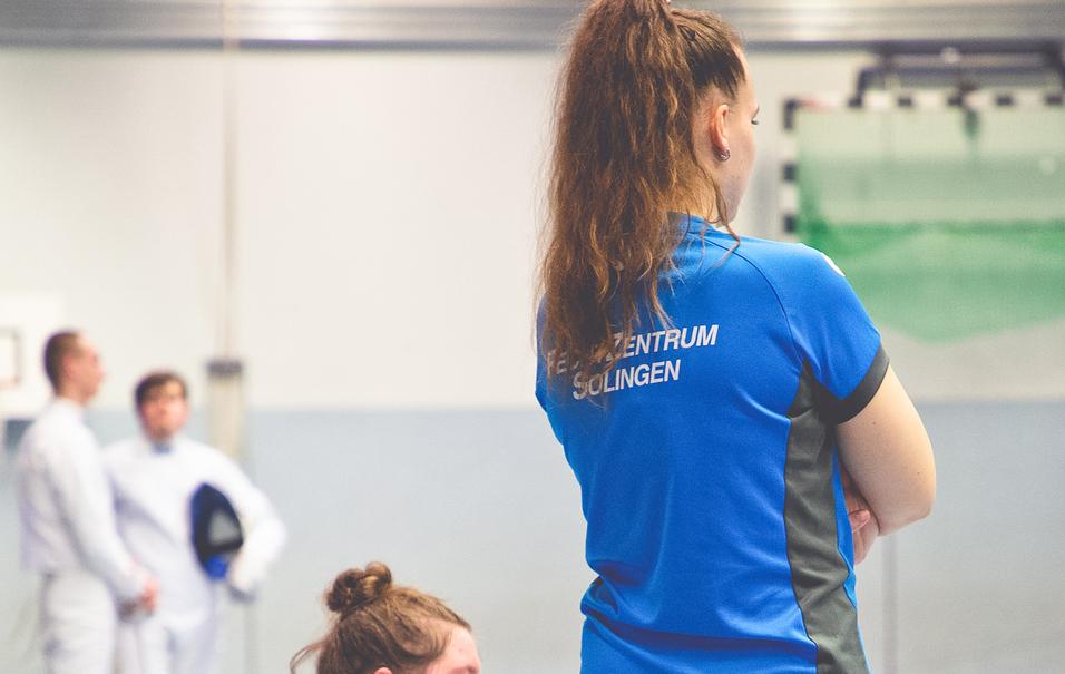 Zwei Personen im Trainingsanzug stehen in einer Sporthalle; eine sitzt, die andere steht und blickt in ihre Richtung.