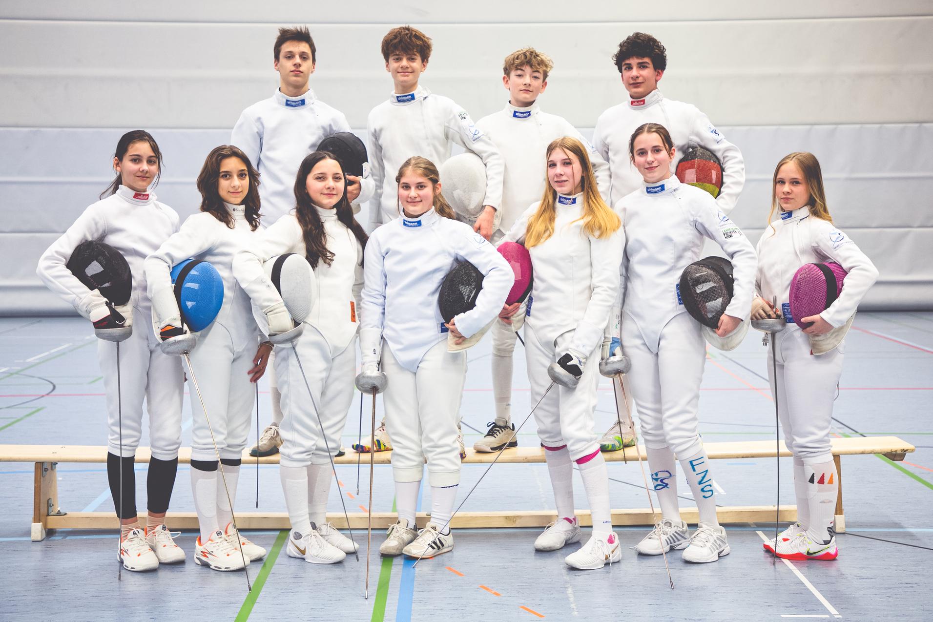 Gruppe von zehn jungen Fechtern in wei&szlig;en Anz&uuml;gen, ausgestattet mit Masken und Degen, in einer Sporthalle.