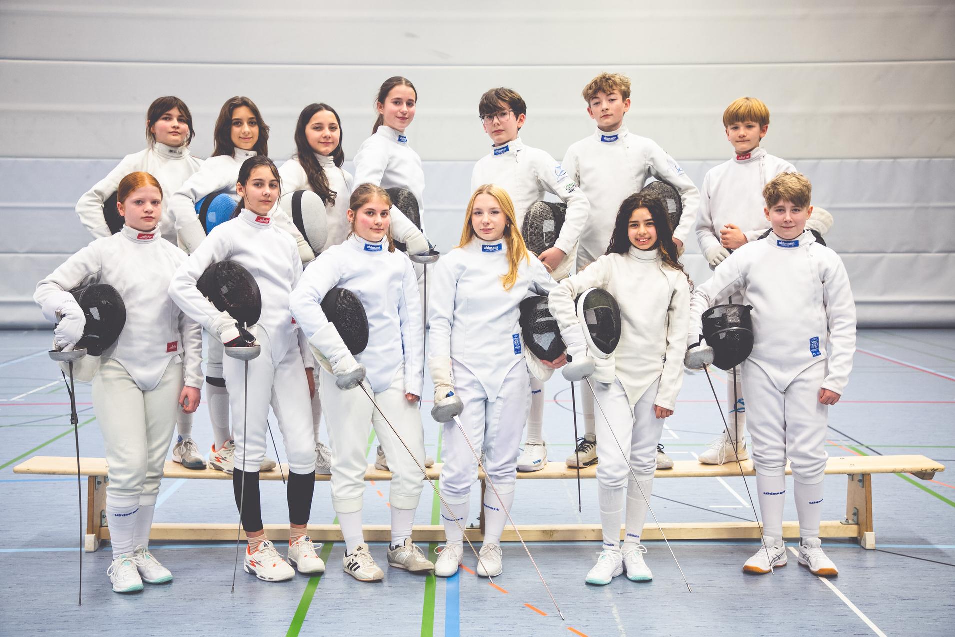 Gruppenfoto von zw&ouml;lf jungen Fechtern in wei&szlig;en Anz&uuml;gen, einige halten Masken und Fechtwaffen.