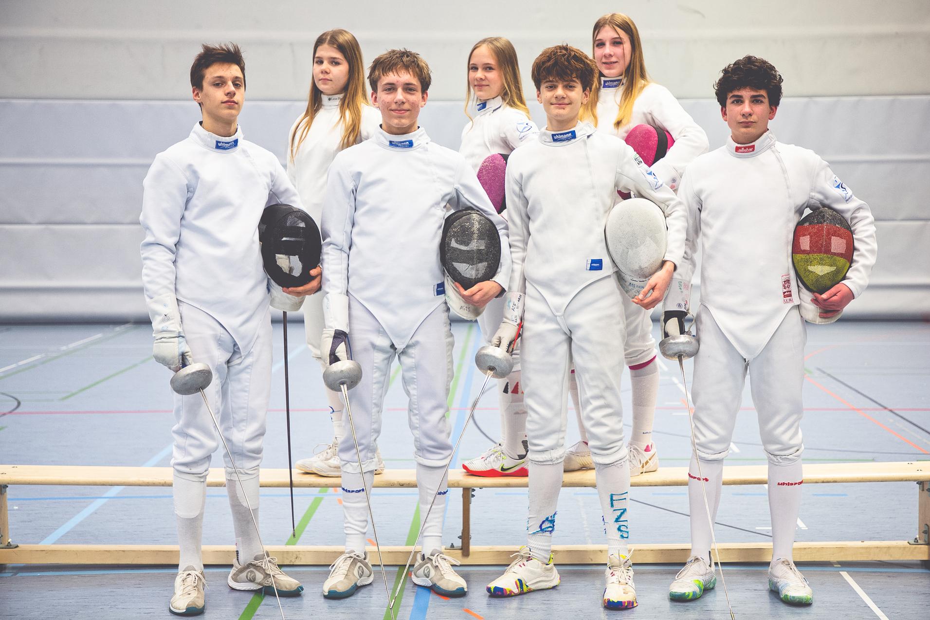Sechs junge Fechter in Wettkampfkleidung posieren mit ihren Degen in einer Sporthalle.