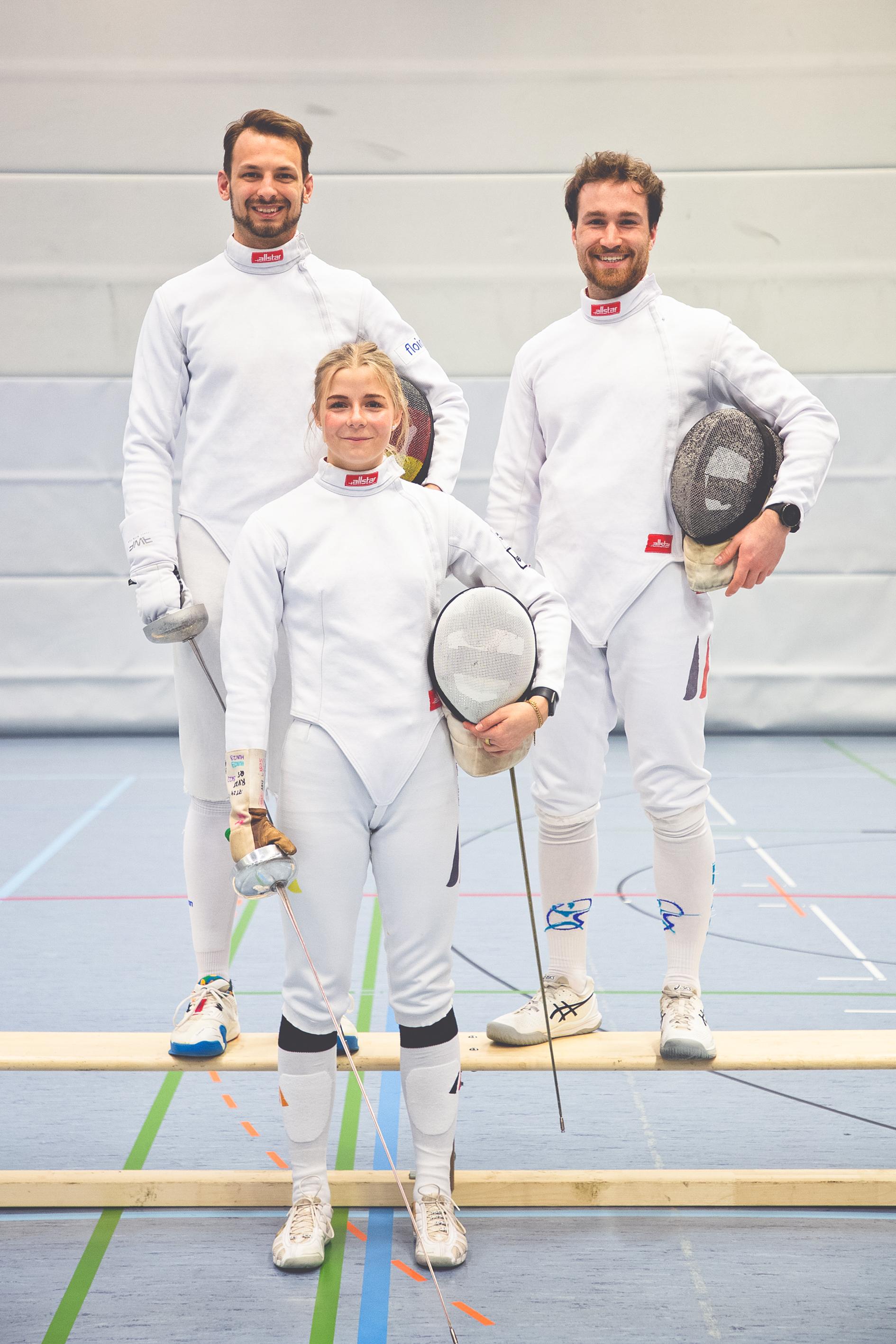 Drei Fechter in Wei&szlig; posieren auf einer Trainingsfl&auml;che, jeder h&auml;lt eine Fechtwaffe und hat eine Maske in der Hand.