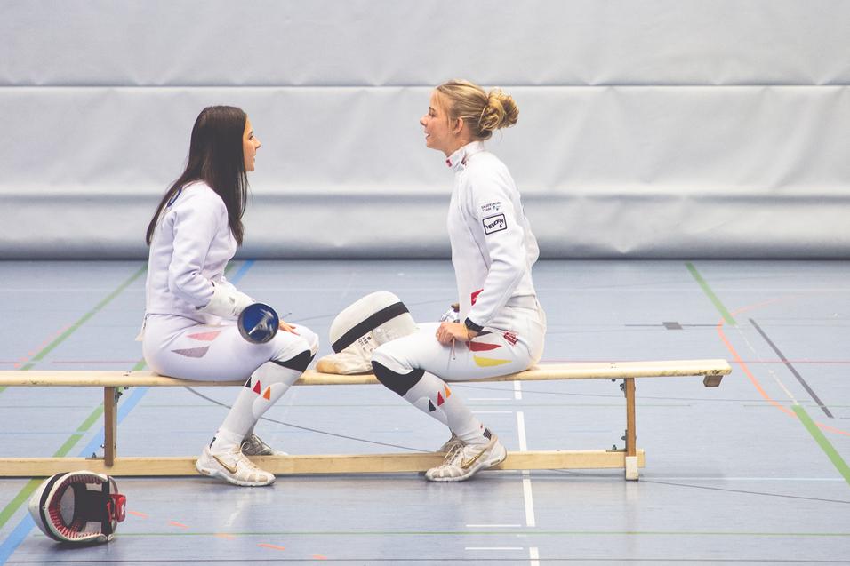 Zwei Fechterinnen sitzen auf einer Bank in einer Sporthalle und führen ein angeregtes Gespräch.