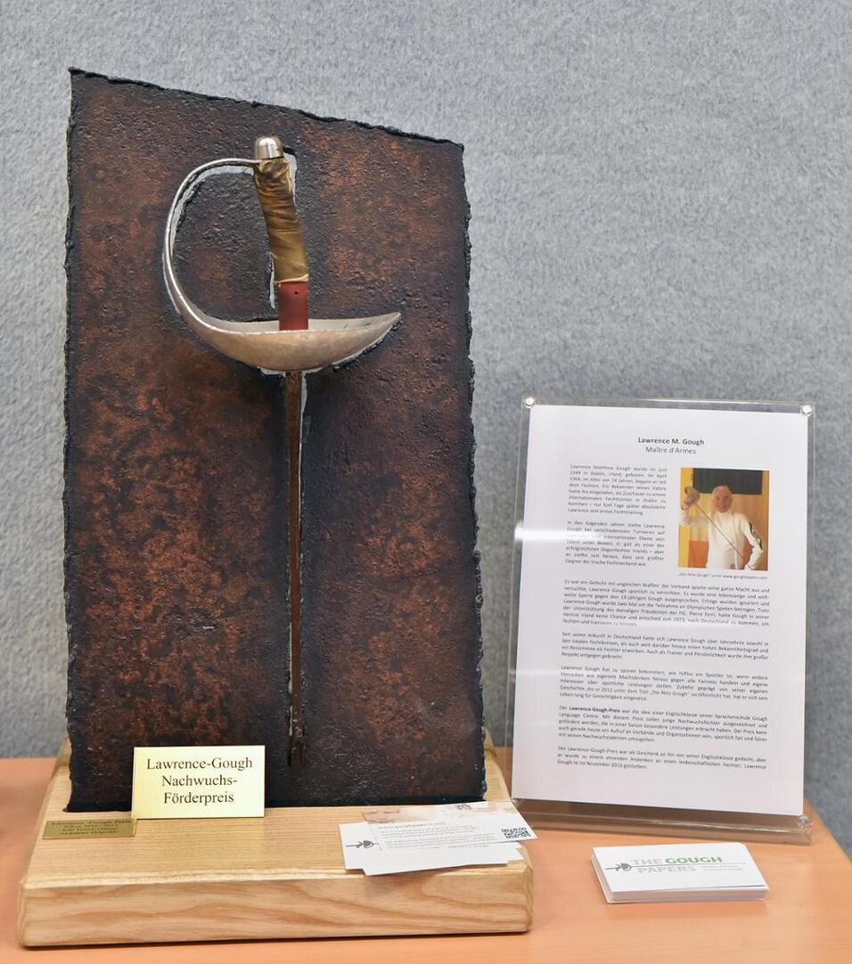Skulptur eines Degen auf einer rustikalen Tafel, daneben Informationen zum Lawrence-Gough Nachwuchspreis.