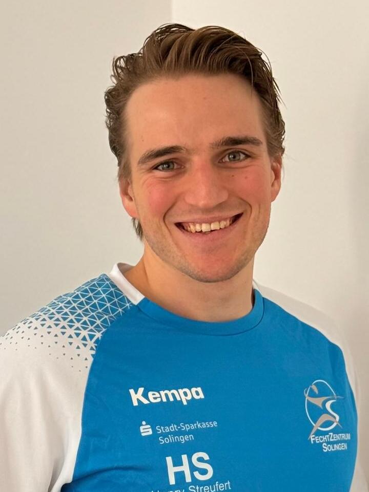 Ein junger Mann l&auml;chelt in einem blauen Sporttrikot mit wei&szlig;en &Auml;rmeln vor einem hellen Hintergrund.