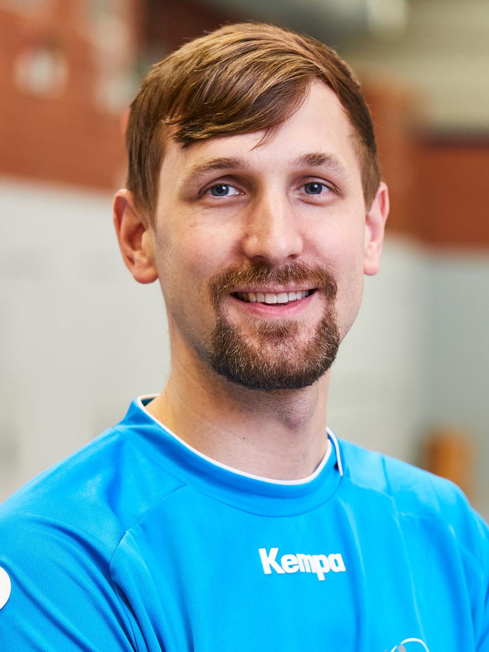 L&auml;chelnder junger Mann mit kurzem Bart in einem blauen Sporttrikot, Hintergrund einer Sporthalle.