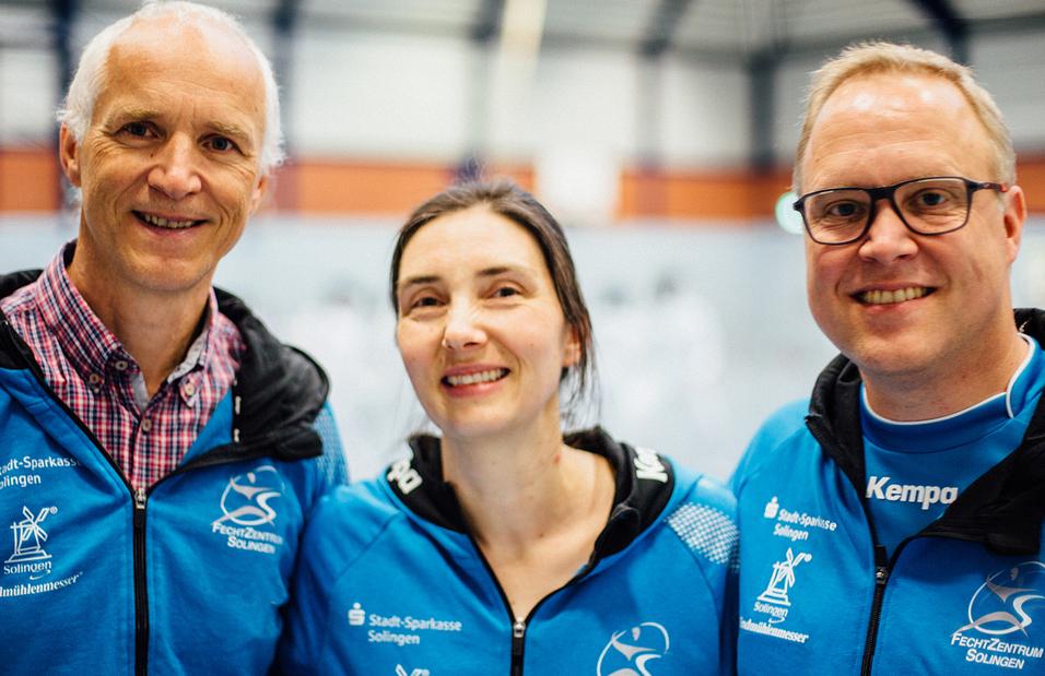 Drei Personen in blauen Trainingsjacken stehen l&auml;chelnd vor einer Turnhalle, im Hintergrund ist eine Sportveranstaltung sichtbar.