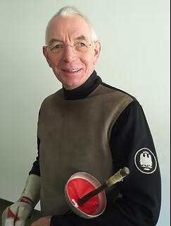 Portrait eines älteren Mannes in Fechtkleidung mit einer Sportmaske und einem Degen in der Hand.
