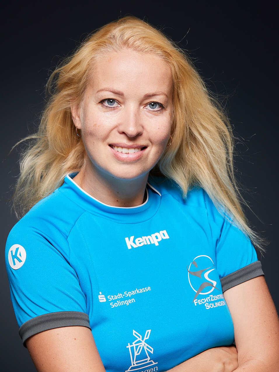 Portr&auml;t einer l&auml;chelnden Sportlerin in blauem Trikot, mit verschr&auml;nkten Armen vor dunklem Hintergrund.