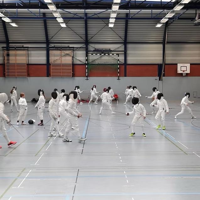 Gruppentraining von Fechtern in wei&szlig;en Anz&uuml;gen in einer Sporthalle, einige f&uuml;hren Angriffe aus, andere warten.