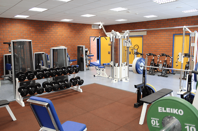 Fitnessstudio mit Gewichten, Kraftgeräten und Trainingsbänken auf einem Gummiboden in einem gut beleuchteten Raum.