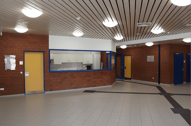 Offene Lobby mit roten Backsteinwänden, weißen Decken und mehreren Türen, inklusive einer Küche im Hintergrund.