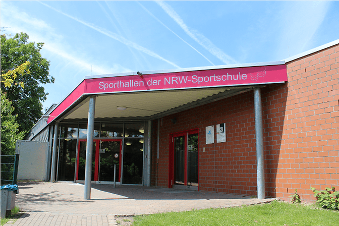 Eingang der Sporthallen der NRW-Sportschule mit rotem Schild und großen Fenstern, umgeben von Grünanlagen.