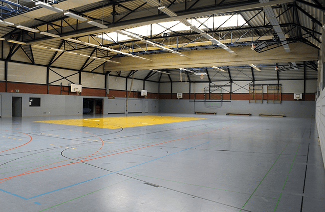 Leere Sporthalle mit gelbem Basketballfeld, mehreren Basketballkörben und grauem Boden mit farbigen Linien.