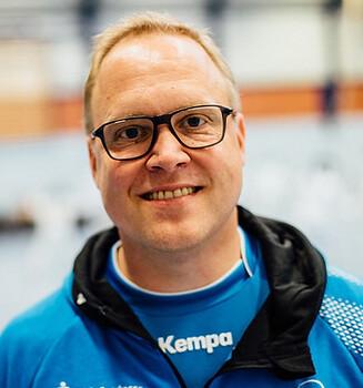 L&auml;chelnder Mann in blauer Sportjacke, hinter ihm eine Sporthalle mit Fechtger&auml;ten sichtbar.