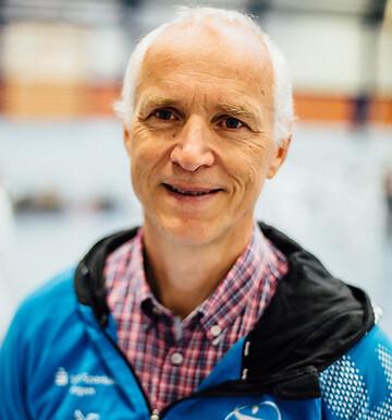 Portr&auml;t eines Mannes in einem blauen Trainingsanzug, l&auml;chelnd in einer Sporthalle mit verschwommenem Hintergrund.