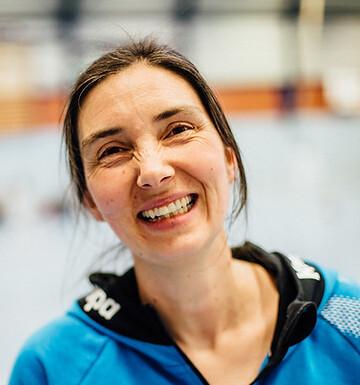 L&auml;chelnde Frau mit langen Haaren in blauer Sportjacke, Hintergrund zeigt eine Sporthalle.