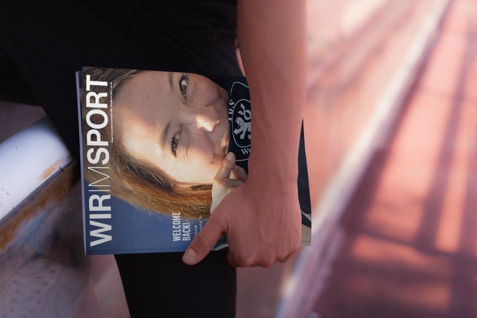 Nahaufnahme einer Person, die ein Magazin mit dem Titel &bdquo;WIR IM SPORT&ldquo; in der Hand h&auml;lt, auf einer Sportanlage sitzend.