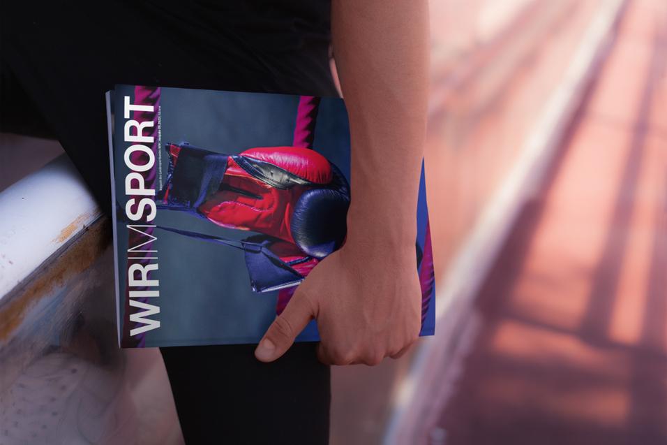 Nahaufnahme einer Hand, die ein Magazin mit dem Titel &bdquo;WIR IM SPORT&ldquo; h&auml;lt, w&auml;hrend sie auf einer Sportbahn sitzt.